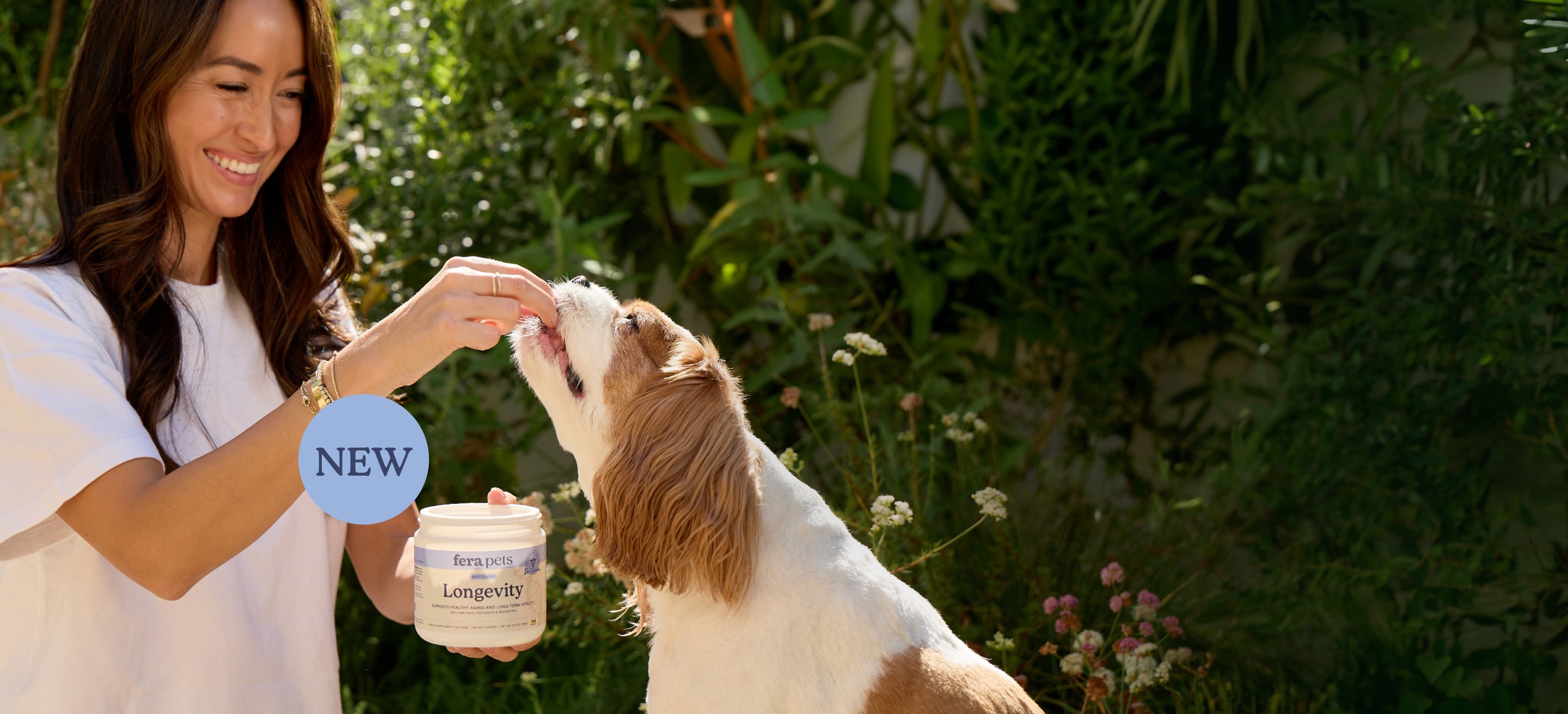 A woman in a white shirt feeds a brown and white dog while holding a jar of Fera Pets pet supplements in a garden setting. A blue circular badge with the word 'NEW' is overlaid on the left side.
